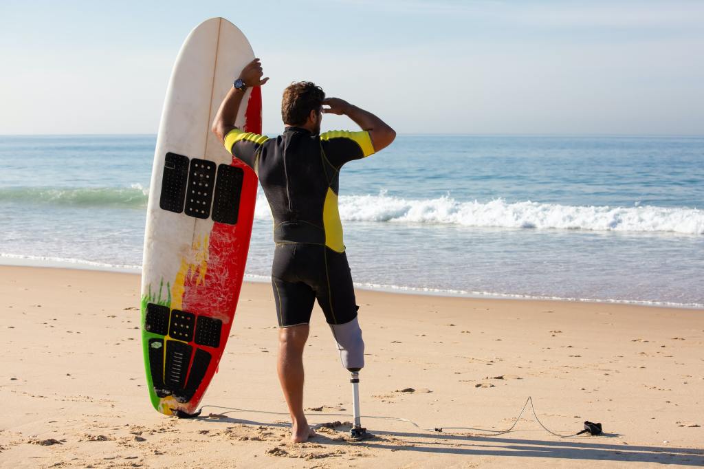 Surfer mit Board und Beinprothese steht am Meer- Perspektivenwechsel mit Prothesen